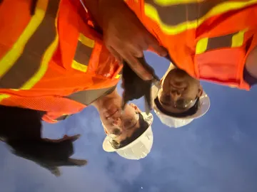 Matteo and a coworker in hard hats and high-vis vests, shot from below against the sky